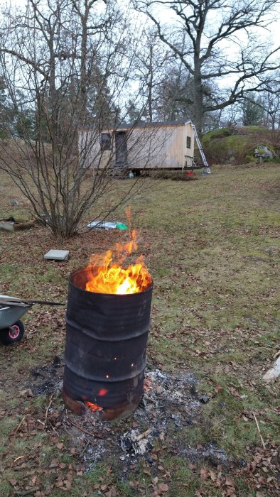 Brinnande trumma med gammalt trä i en trädgård, med byggprojekt i bakgrunden.