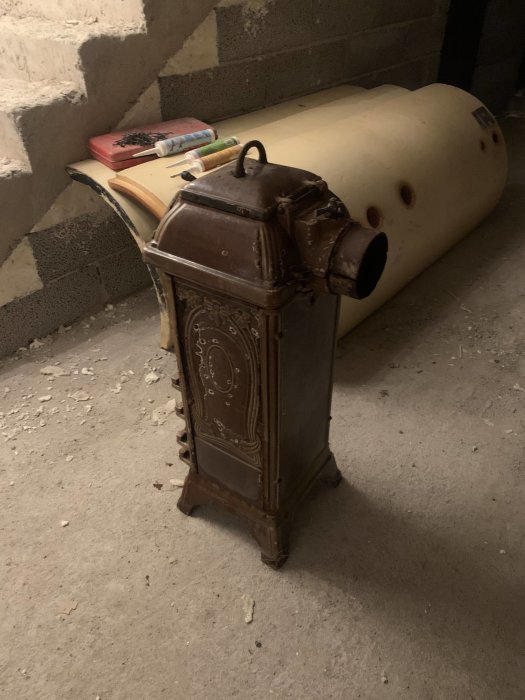 Gammal gjutjärnskamin med rost och skador bredvid en galvaniserad tank och byggmaterial på golv.