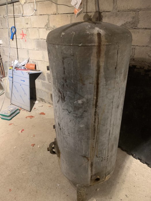 Äldre galvaniserad vattentank med rost och läckagespår, står i ett oinrett utrymme.