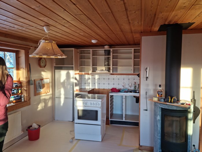Kök under renovering med borttagna skåpluckor och kakel, vedspis i förgrunden och köksspis i bakgrunden.