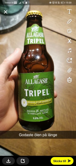 Hand håller en flaska Allagash Tripel öl med texten "godaste ölen på länge" synlig på skärmen.