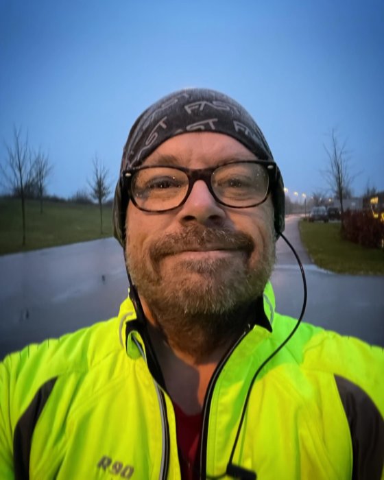 Person med glasögon, bandana och reflexjacka ler mot kameran utomhus i skymningen.