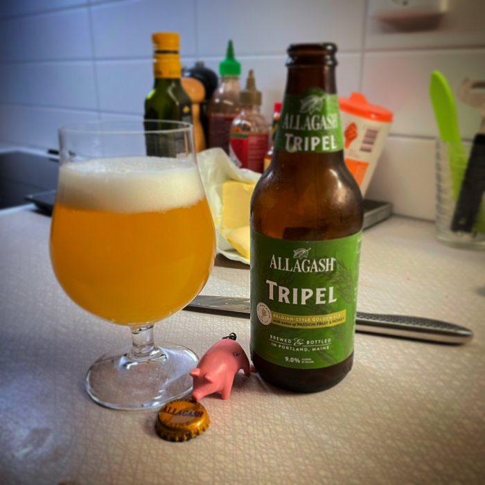 Ölglas med skum topp bredvid en Allagash Tripel ölflaska och en rosa leksaksgris på ett köksbord.