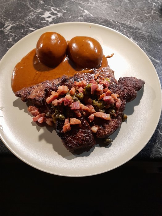 Stekt lever toppad med bacon och kapris med två runda potatisar och gräddsås på en vit tallrik.