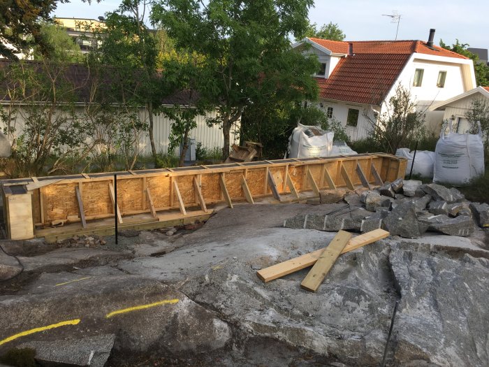 Träform för gjutning av stödmur på granitmark med utspridda stenblock och byggmaterial, vit villa i bakgrunden.