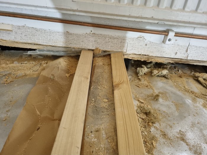 Två träreglar placerade på varsin sida av ett upprivet golv nära en vägg och en radiator.