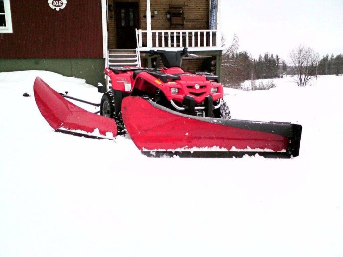 Röd ATV med monterad vikplog framför ett snöigt landskap och hus