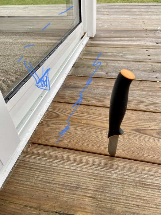 Ett fönster med blå markeringar på glaset och en kniv stående vertikalt på en träterrass.