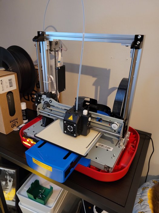 3D-skrivare med blå filamentrulle på en arbetsstation, ej strukturell plast syns.