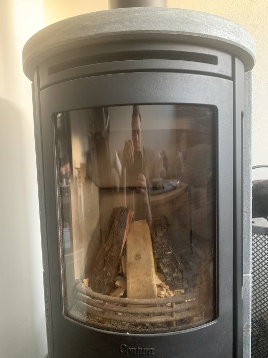 Contura 560T kamin med svängt glas och ved inuti, reflektion av en person som tar bilden.