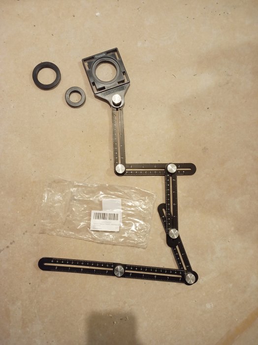 Svart aluminiummätverktyg med sex sidor, skenor och muttrar av metall, och plastdelar, på en ljus yta.