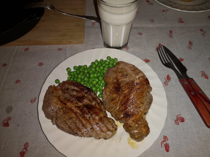 Två grillade fläskkotletter med gröna ärtor på en vit tallrik, bredvid ett glas mjölk och bestick.