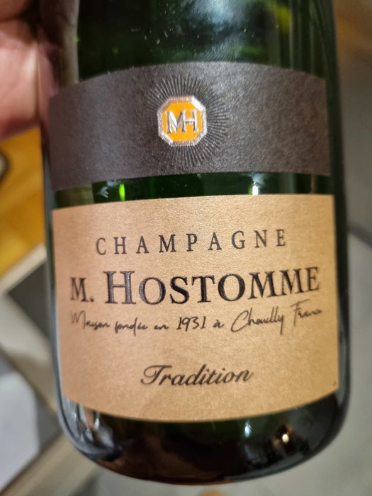 Närbild av en champagneflaska med etiketten "M. HOSTOMME Tradition" och texten "Maison fondée en 1931 à Chouilly - Epernay".