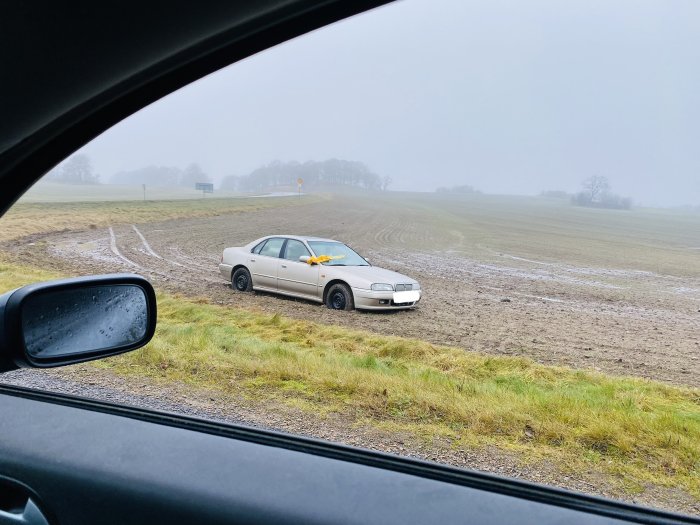 Bil fastkörd i lera vid en åker, sedd från insidan av ett annat fordon, dimmig dag.