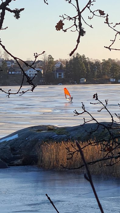 En person isseglar på en frusen sjö vid solnedgång med träd och hus i bakgrunden.