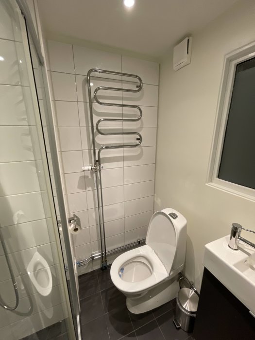 Handdukstork i form av böjda rör på en badrumsvägg ovanför en toalett, bredvid en dusch och ett handfat.