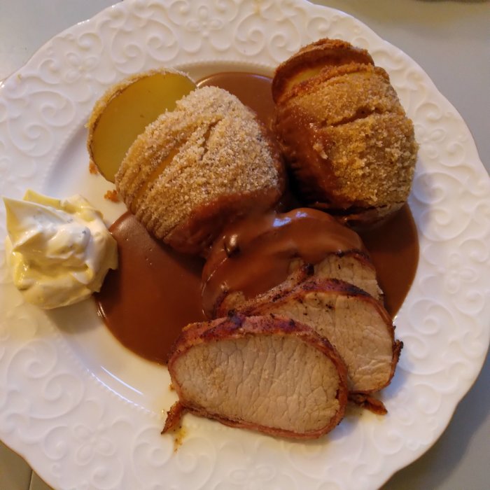 Baconlindad kotlettrad och hasselbackspotatis med skal, gräddsås och klickar av bearnaisesås på vit tallrik.