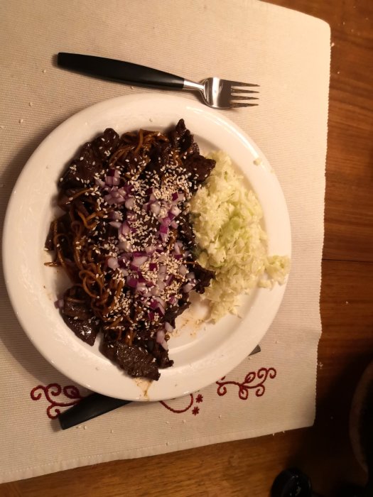 Wokad blandning av nudlar och kött toppad med hackad röd lök och sesamfrön, serverad med vitkålssallad.