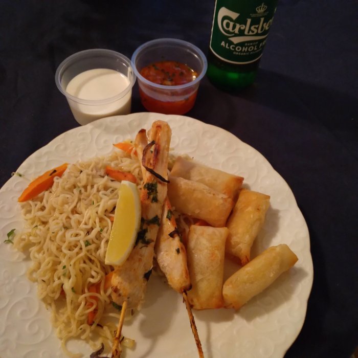 Tallrik med wokade nudlar, marinerade kycklingspett, vårrullar, aioli, sötsur sås och en alkoholfri Carlsberg.