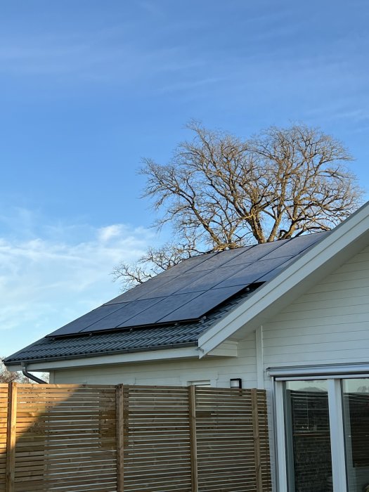 Solpaneler installerade på taket av ett hus med träfasad, under en blå himmel och grenar från ett träd överst.