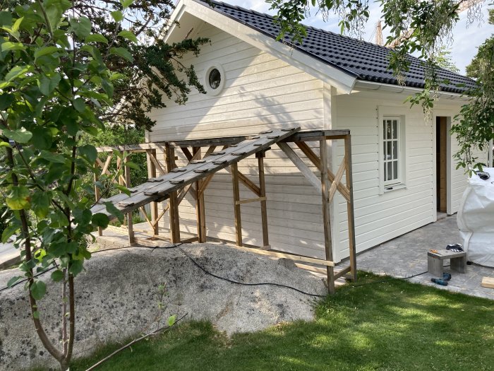 Pågående Attefallsbygge med färdigställd vit fasad, vindduk, installerade fönster och halvfärdig terrasskonstruktion.