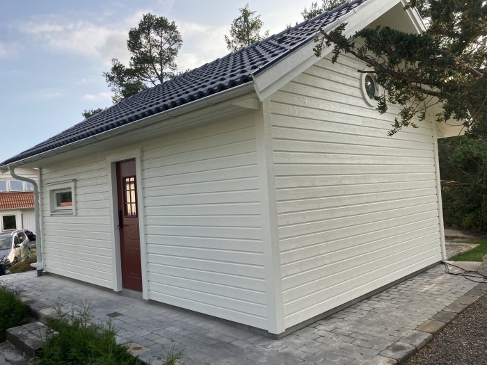Nybyggt hus med vit panel, röd dörr och stenlagd uppfart, klar exteriör efter renovering.