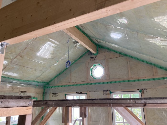 Inre av ett hus under renovering med isolering, träbjälkar och synlig rördragning i taket.