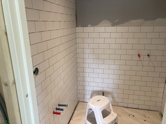 Hörn av ett badrum under renovering med vita kakelväggar och spacers, ej fogade, och en plastpall.