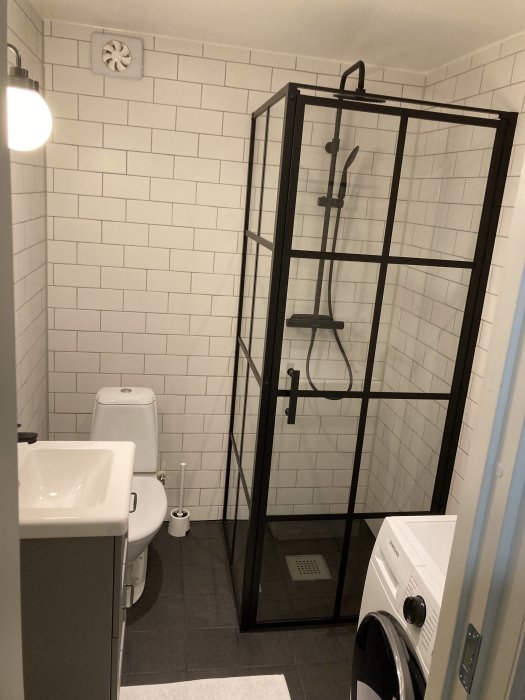 Renoverat badrum med vita kakelväggar, svart duschutrymme, toalett och tvättmaskin.