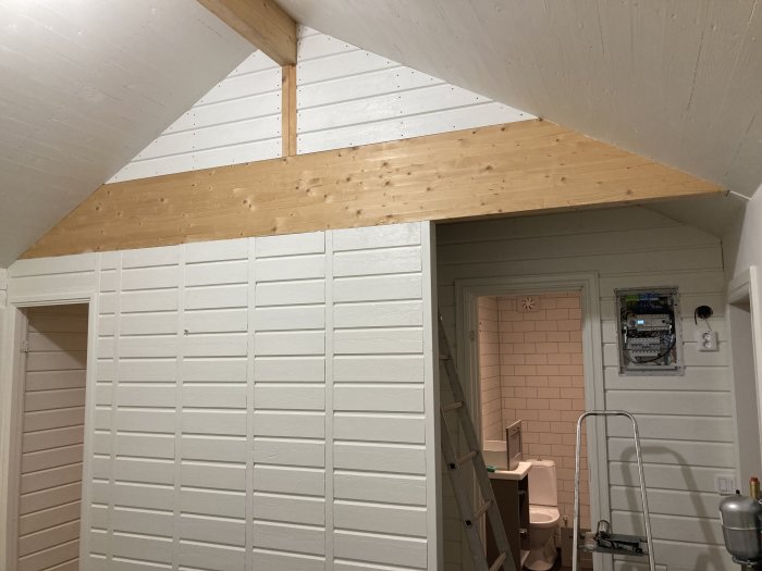 Renoverat rum med vitmålade väggar och ett obehandlat träloft, stege intill en dörröppning, samt elskåp och badrum i bakgrunden.