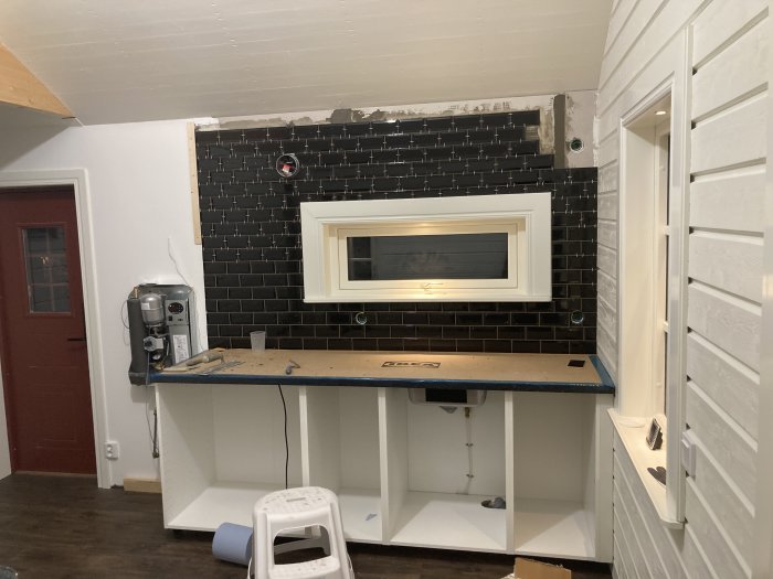 Kök under renovering med svarta kakelplattor ovanför vit inredning och verktyg på bänken.