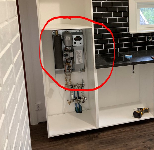 Köksskåp öppet med en mini elpanna för golvvärme installerad, svarta kakelplattor i bakgrunden.