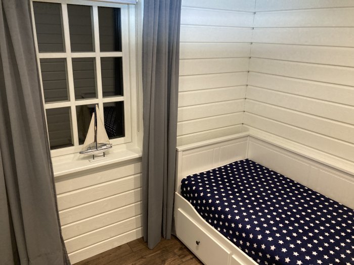 Hörn av ett sovrum med vita panelväggar, fönster med dekorationsskepp och en säng med stjärnmönstrat överkast.