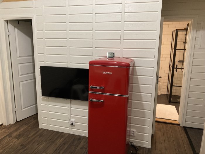 Renoverat kök med röd retro kylskåp, vit Z-panel på väggar och mörkt trägolv, utsikt mot dörröppning till badrum.