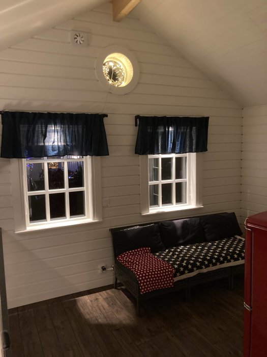 Vitpanelat rum med mörka träklinkers, två vitmålade fönster med mörka gardiner och svart soffa med stjärnmönstrade kuddar.