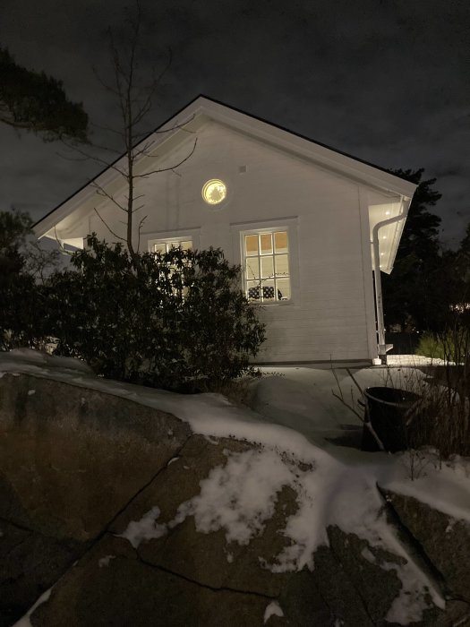 Ett nymålat hus på kvällen med belysning och snö på marken, synlig genom grenar och buskar.