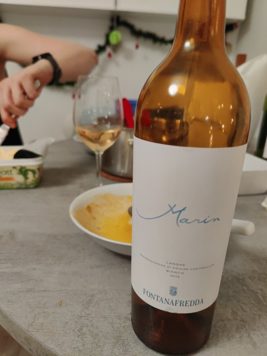 Flaska av Fontanafredda 2016 Langhe Bianco öppnad vid matbord, med ett glas vin och suddig fiskgryta i bakgrunden.
