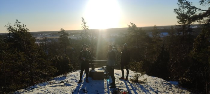 Personer står på en snötäckt utsiktsplats i motljus med en solnedgång i bakgrunden.