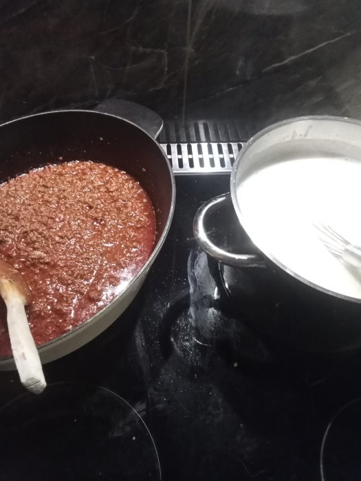 Ingredienser till lasagne med köttfärssås i stekpanna och bechamelsås i kastrull på spis.