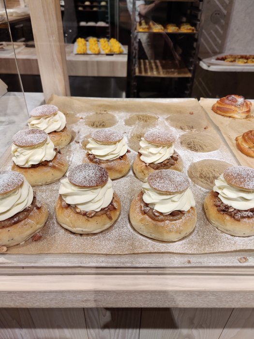 Flera nybakade semlor med grädde och mandelmassa på ett bagerifat, bakgrund av bakverk.