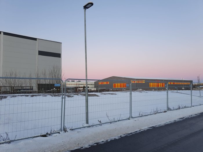 Industribyggnader vid gryning med snötäckt mark och ett metallstängsel i förgrunden.