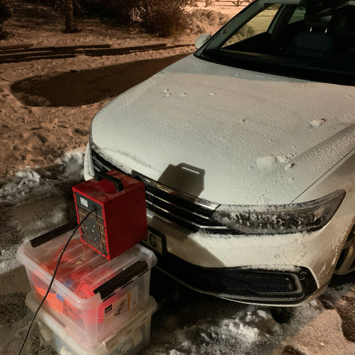 Bil täckt med frost och snö framför hus, med laddningsbox kopplad till fronten.