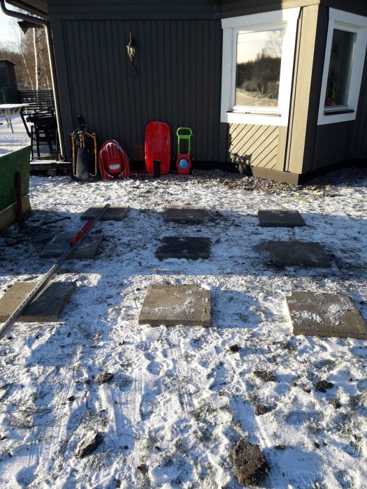 Betongplattor på mark täckt med snö, placerade med mellanrum, nära en husvägg med fönster och leksaker.