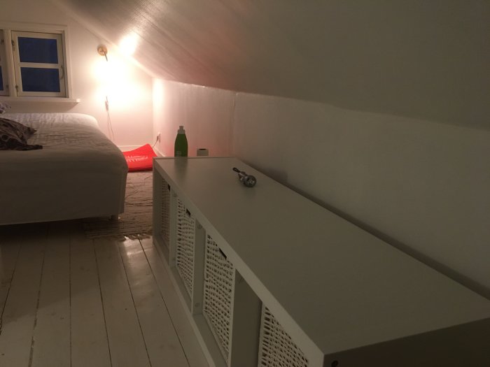 Del av ett vitmålat vindrum med snedtak, säng, sidobord och inbyggd förvaring.