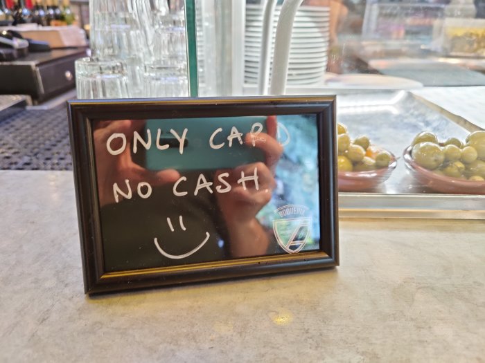 Skylt med texten "ONLY CARD NO CASH" och en leende smiley framför en bar med glas och oliver.