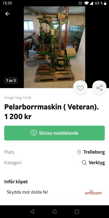 Grön veteran pelarborrmaskin i ett garage med ett rött verktyg och en gammal grönmålad släpvagn i bakgrunden.