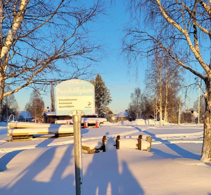 Vinterlandskap med snö, skylt som säger "Pusshållplats", soligt väder och träd i bakgrunden.