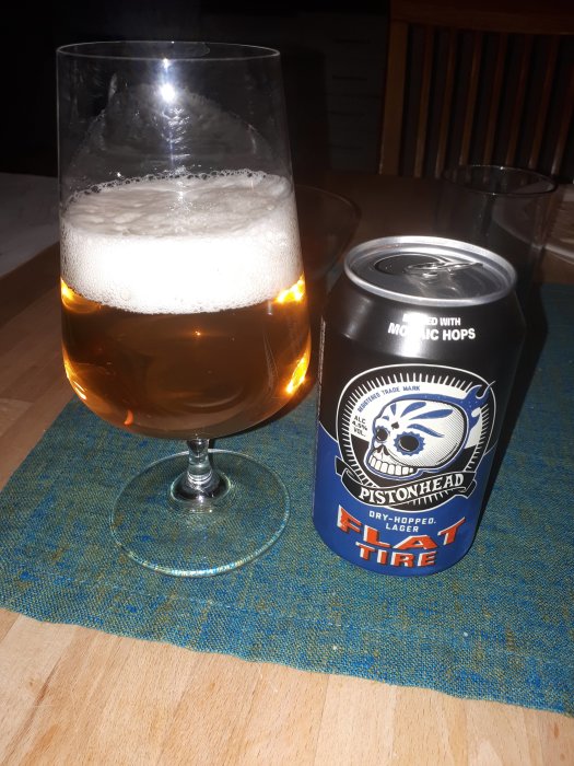 Ölglas fyllt med ljus öl bredvid en burk Pistonhead Flat Tire torrhumlad lager på blå duk.