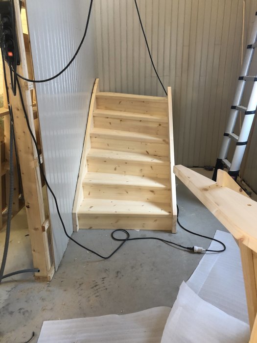 Trätrappa under konstruktion i verkstadslokal med synliga elledningar och verktyg på betonggolv.