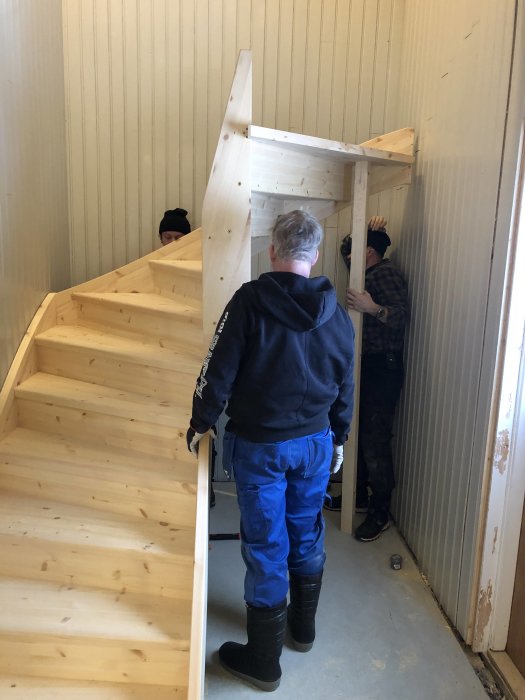 Två personer monterar en trätrappa inuti ett hus, med de ofärdiga väggarna synliga.